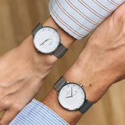Two crossed wrists in striped shirts, each wearing a white-dial watch on a mesh strap.