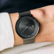 Gunmetal watch and mesh strap on a wrist with a white knit sleeve, gray dial in view.