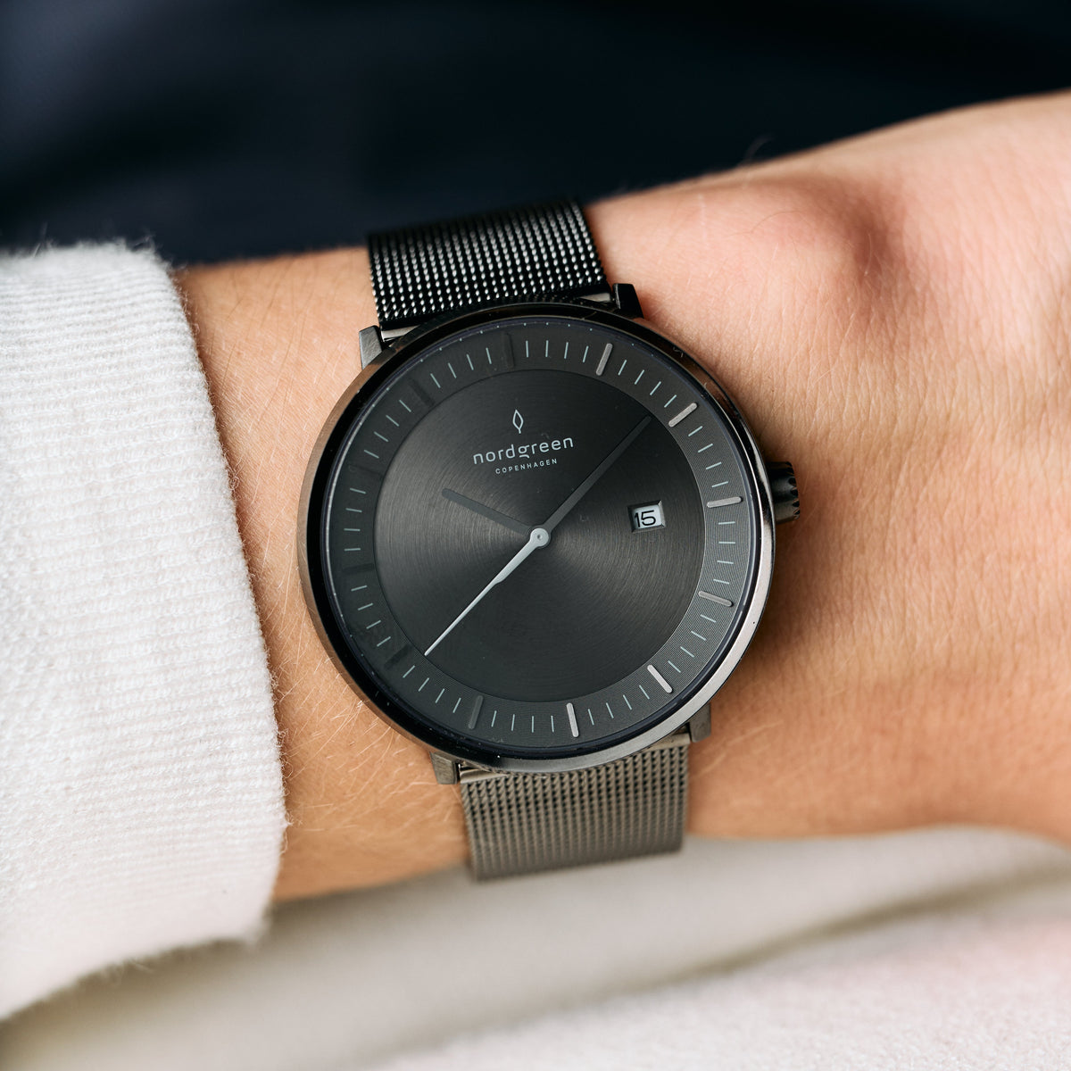 Gunmetal watch and mesh strap on a wrist with a white knit sleeve, gray dial in view.