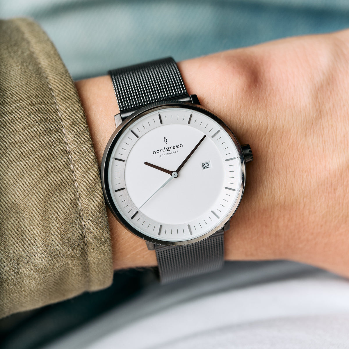 White dial in a gunmetal case on a dark mesh strap, worn under an olive jacket sleeve.