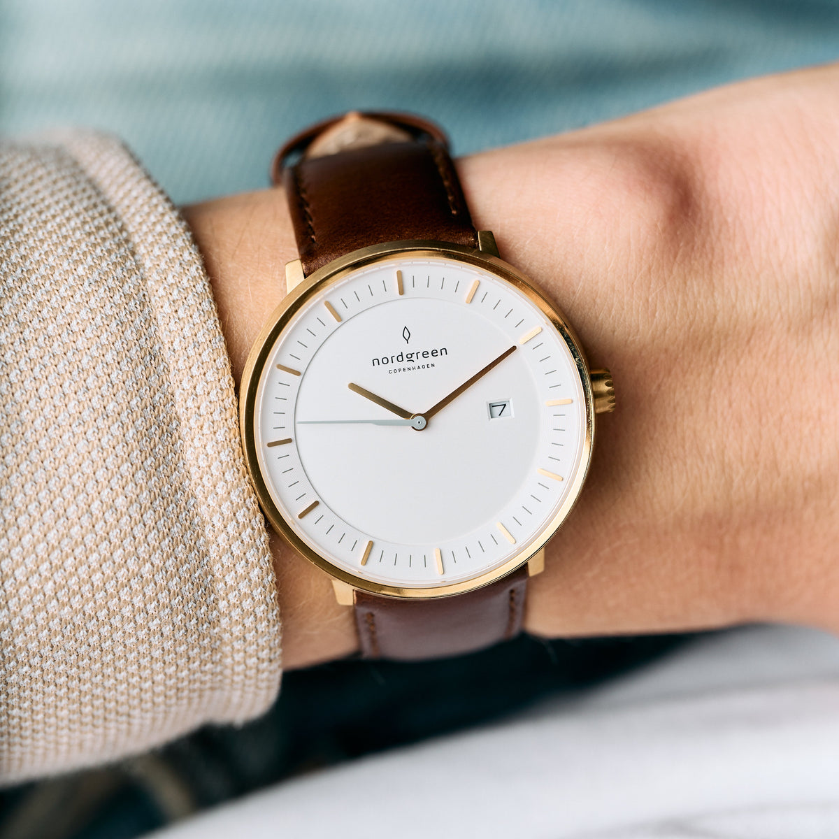 Gold-tone case and white dial on a brown strap, worn with a beige knit sleeve.