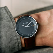 Worn on a wrist under a gray-green shirt cuff, black dial with orange accent.