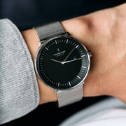 Silver-tone watch with a black dial on a mesh strap, worn with a gray sweater.