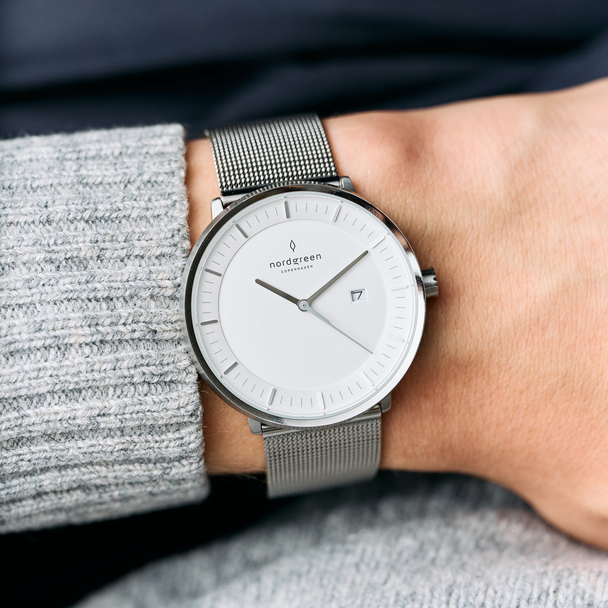 A gray knit sleeve pulled back to show the white dial and silver-tone mesh strap.