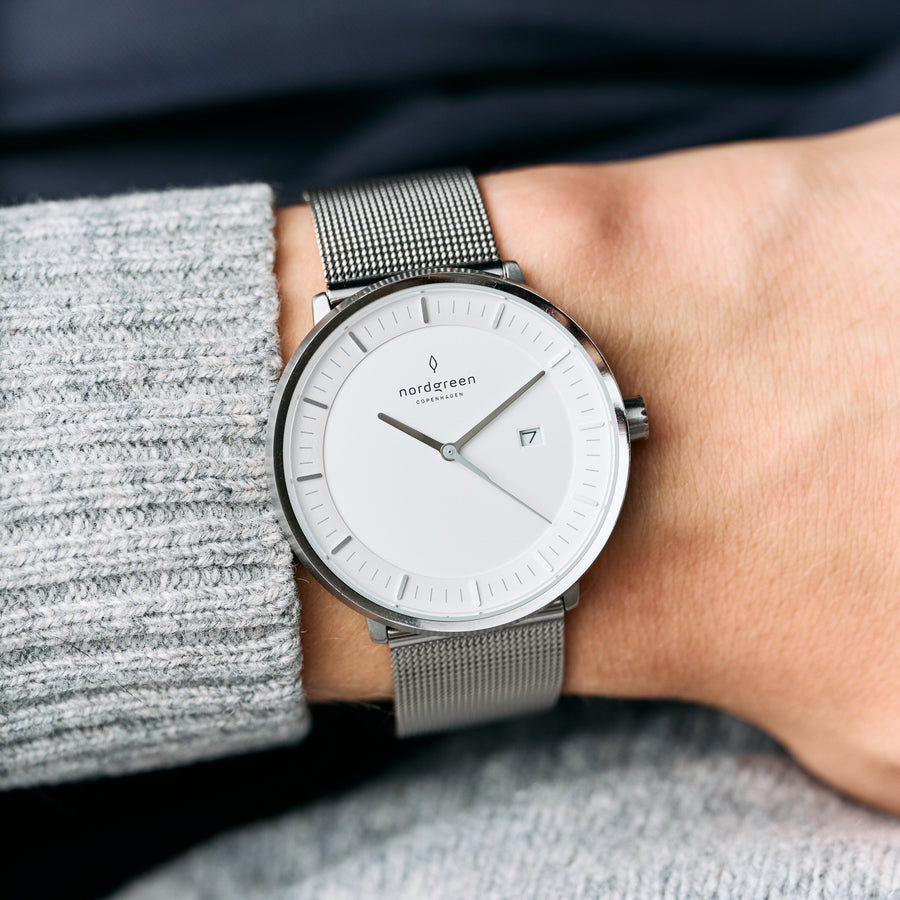 A gray knit sleeve pulled back to show the white dial and silver-tone mesh strap.