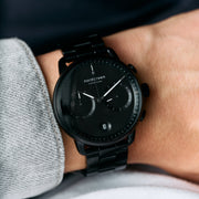 The black bracelet watch worn on a wrist beside a gray sleeve.