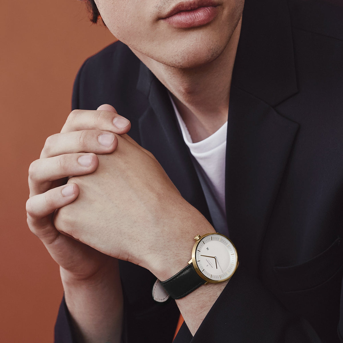 A man in a dark blazer with hands clasped, wearing the gold-tone watch.