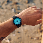 Wrist shot of the black and turquoise watch outdoors over sand.