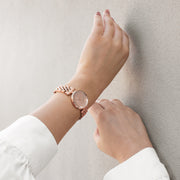 Slim rose gold watch on a wrist resting against a pale wall.