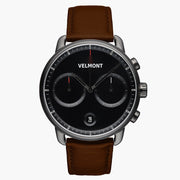 Chronograph-style watch with a black dial and gunmetal case on a brown strap.