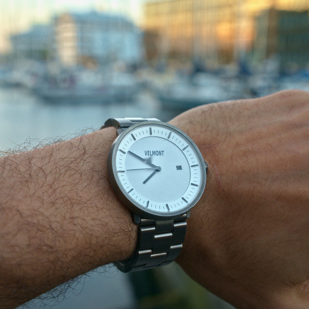 Worn on a wrist by the harbor, white dial and gunmetal bracelet in evening light.