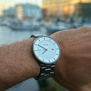 Gunmetal watch on a wrist with blurred harbor buildings behind.
