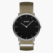 Black-dial watch with a silver-tone case on a khaki woven strap, front view.