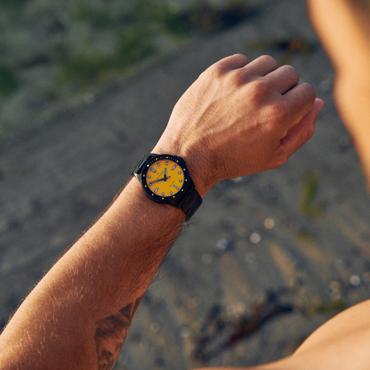 Watch with a yellow dial worn outdoors on a raised wrist over rocky ground.